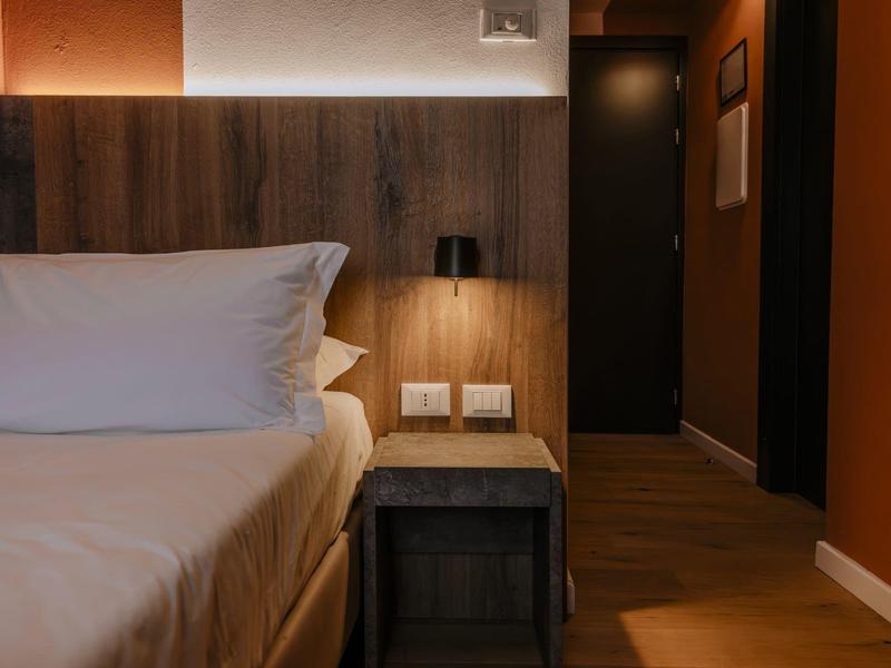 Moderní hotelový pokoj s bílou postelí, dřevěným panelem a nočním stolkem s lampou.