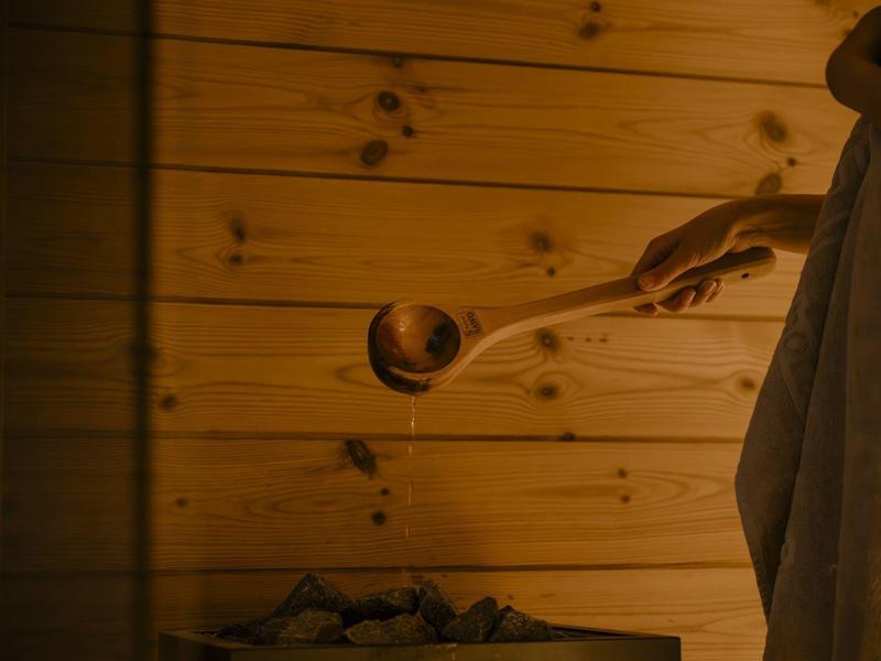 Osoba nalévá vodu dřevěnou naběračkou na kameny v sauně