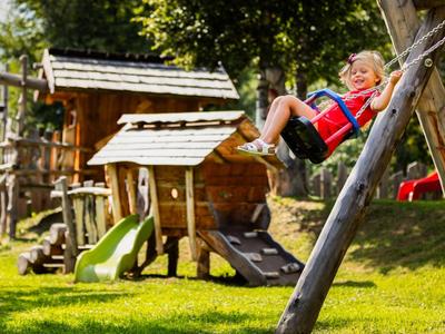 Enfant souriant sur une balançoire dans un jardin ensoleillé avec aire de jeux et cabanes en bois.