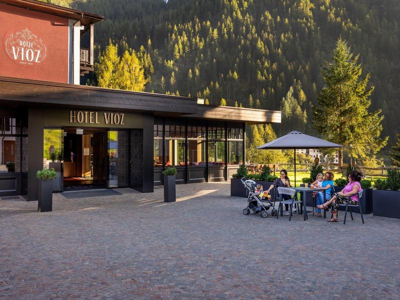 Hôtel avec terrasse et clients assis sur des chaises confortables, entouré de montagnes boisées sous le soleil.