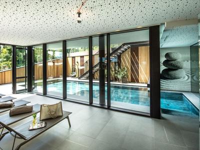 Modern binnenzwembad met ligstoelen en grote glazen wanden met uitzicht op de buitenruimte.