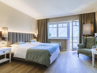 Chambre d'hôtel confortable avec lit double, fauteuil, tables de chevet et grande fenêtre avec rideaux.