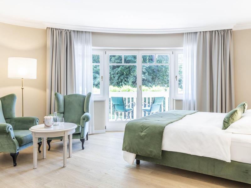 Chambre d'hôtel lumineuse avec lit double, fauteuils verts et vue sur terrasse par de grandes fenêtres.