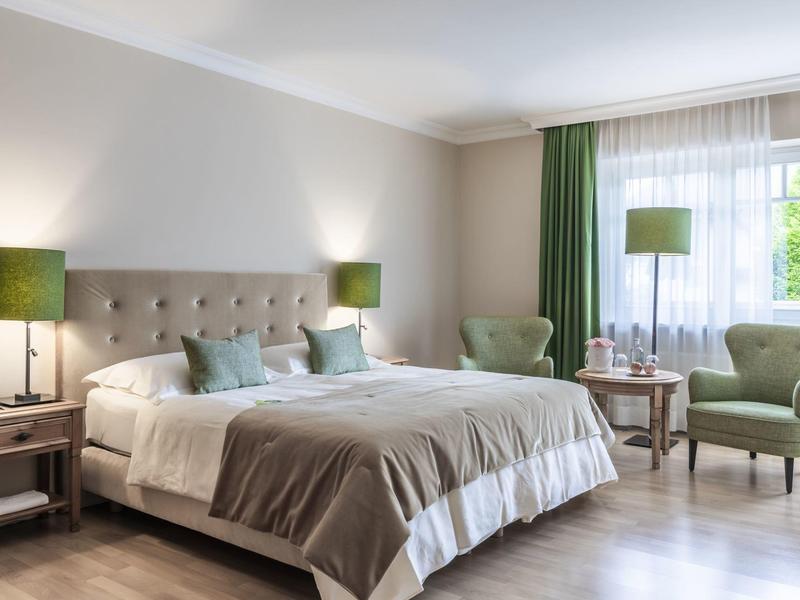 Chambre d'hôtel lumineuse avec un grand lit, des rideaux verts et deux fauteuils près de la fenêtre.