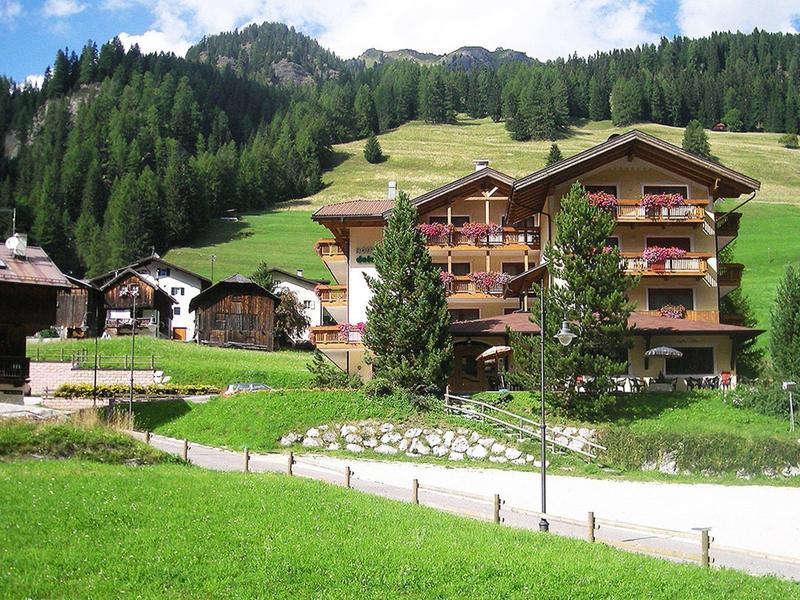 Großes Chalet-Hotel vor bewaldetem Berghang und grünem Wiesenfeld bei klarem Himmel.
