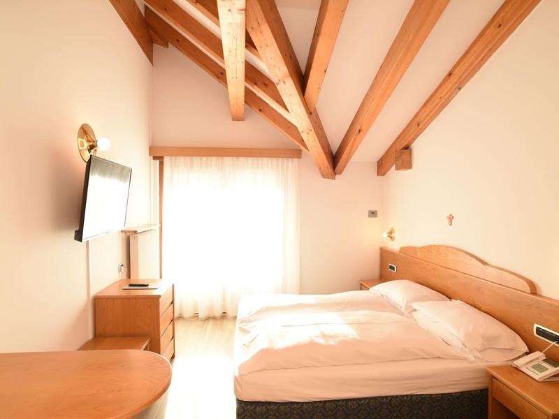 Jasna sypialnia z drewnianymi belkami, podwójnym łóżkiem, stolikami nocnymi i telewizorem na ścianie.