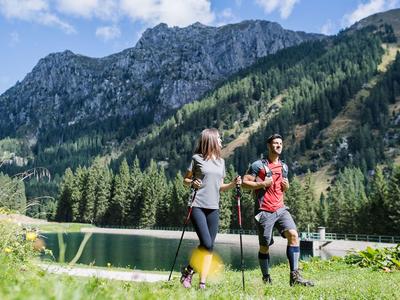 Deux randonneurs avec des bâtons au bord d'un lac devant des montagnes vertes par une journée ensoleillée.