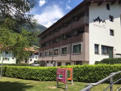 Dreistöckiges Hotelgebäude mit Balkonen vor grüner Hecke und Bäumen unter blauem Himmel.