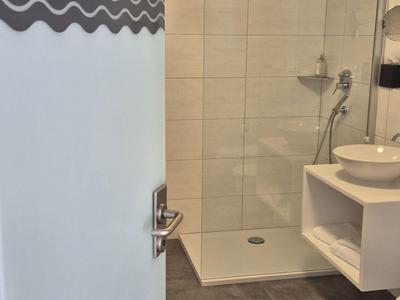 Blick durch eine halb geöffnete Tür auf eine moderne, helle Hotelbadezimmer-Dusche mit weißem Waschbecken.