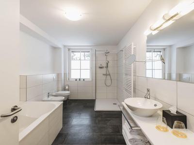 Helles Badezimmer mit Badewanne, Dusche, Waschbecken und Fenster mit weißem Design.
