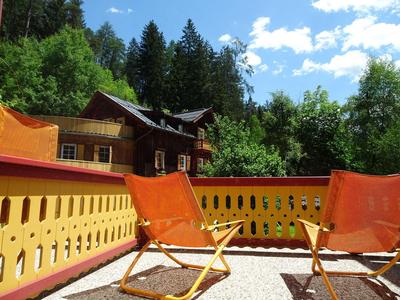 Zwei orange Liegestühle auf einer Terrasse mit gelbem Geländer umgeben von Bäumen bei sonnigem Himmel.