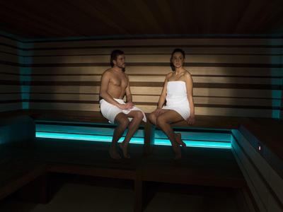 Un couple assis en serviettes sur un banc dans un sauna moderne avec éclairage bleu.