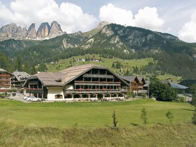 Hotel in alpiner Landschaft mit grünen Wiesen und Bergen im Hintergrund unter bewölktem Himmel.