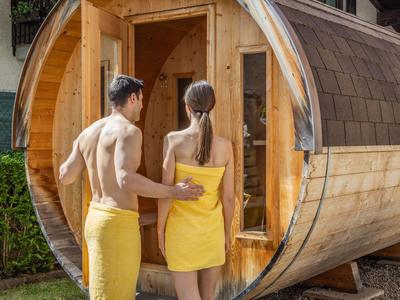 Zwei Personen in gelben Handtüchern vor einer runden Holzsauna im Freien.