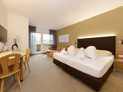 Modernes Hotelzimmer mit großem Bett, Schreibtisch, Fernseher und Sitzbereich in warmen Farbtönen.