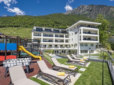 Moderner Hotelkomplex mit Liegestühlen und Kinderspielplatz vor grünen Bergen.