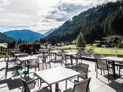 Freiluftterrasse mit Tischen und Stühlen vor bergiger Landschaft bei sonnigem Wetter.