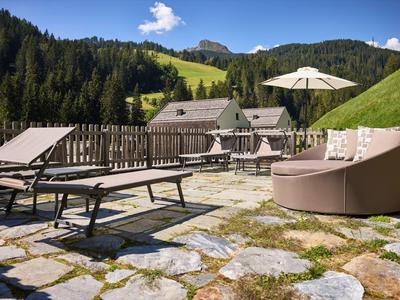 Terrasse mit Liegestühlen und Sitzmöbeln vor einer grünen Berglandschaft unter blauem Himmel.