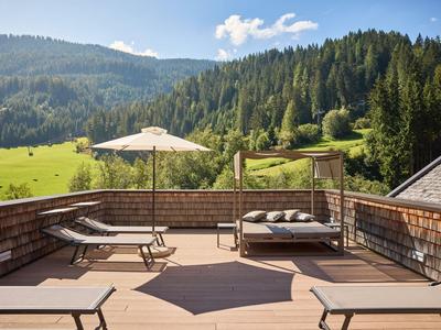 Balkon mit Liegen und Sonnenschirm, umgeben von grünen Hügeln und Wäldern.
