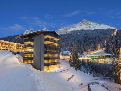 Complesso alberghiero in paesaggio montano innevato al crepuscolo con edifici illuminati.