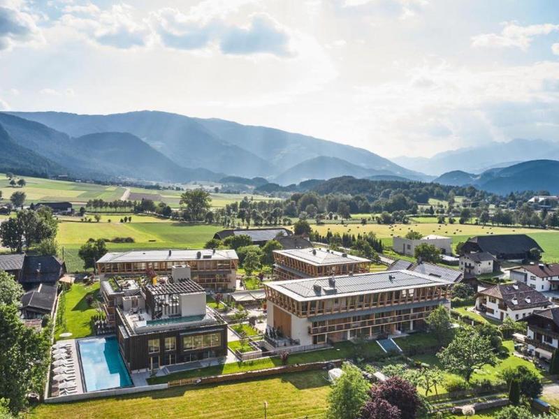 Pohled na hotel s venkovním bazénem, obklopený zelenými poli a horami v pozadí.