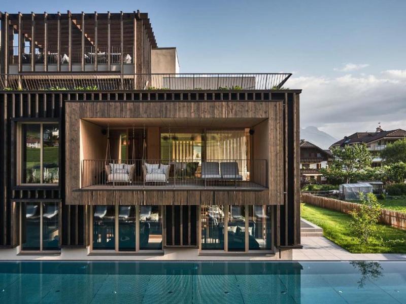 Moderní hotel s černou fasádou, dřevěnou verandou a bazénem v popředí.