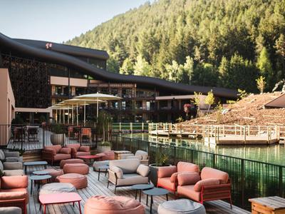 Außenbereich eines Hotels mit gemütlichen Sitzgelegenheiten neben einem Fluss und bewaldeten Hügeln im Hintergrund.