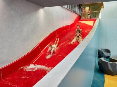 Three children sliding down a red indoor water slide in a modern setting.