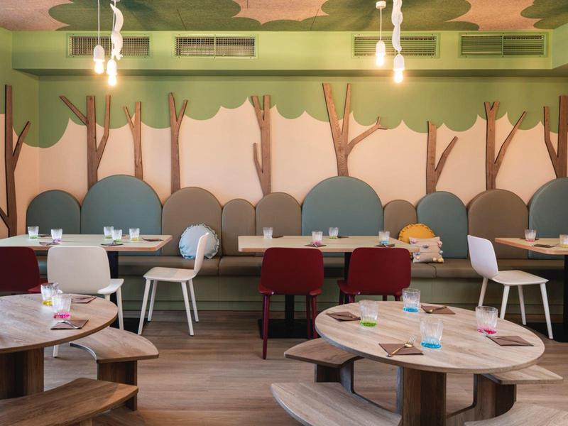 Modern dining area with round wooden tables and upholstered chairs against a green wall with tree design