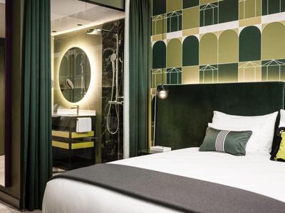 Modernes Hotelzimmer mit großem Bett, grün-gemusterten Wänden und rundem beleuchtetem Spiegel im Bad.