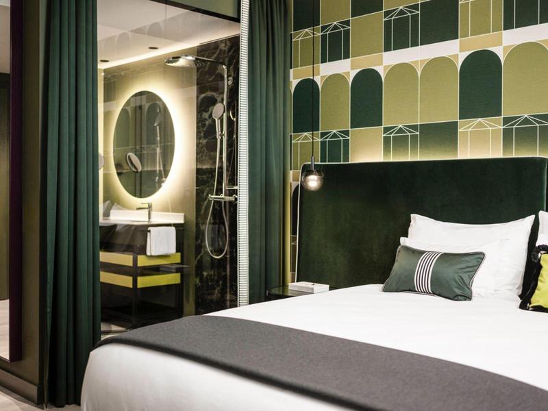 Modernes Schlafzimmer mit grünem Kopfteil, bunten Kissen und einem angrenzenden Badezimmer mit rundem Spiegel.