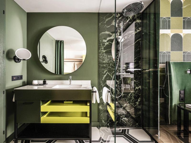 Modernes Badezimmer mit grünen Wänden, rundem Spiegel, gelbem Waschbeckenunterschrank und Glasdusche.