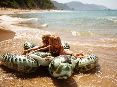 Child playing on an inflatable turtle on a sunny beach shore with waves and mountains in the background