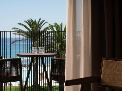 Balcony with table and chairs overlooking palm trees and sea under clear sky.