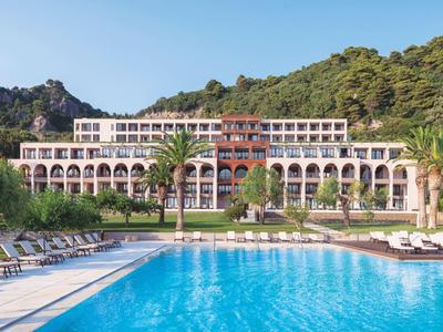 Hôtel de luxe avec colonnade, palmiers et grande piscine devant des collines boisées.