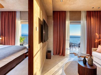 Modern hotel room with bed, TV, and balcony overlooking the sea and lounge area.