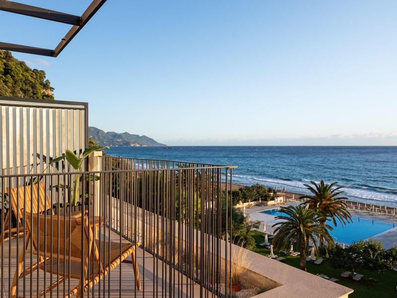 Balcony with chairs overlooking pool, palm trees, and ocean under clear sky at resort.