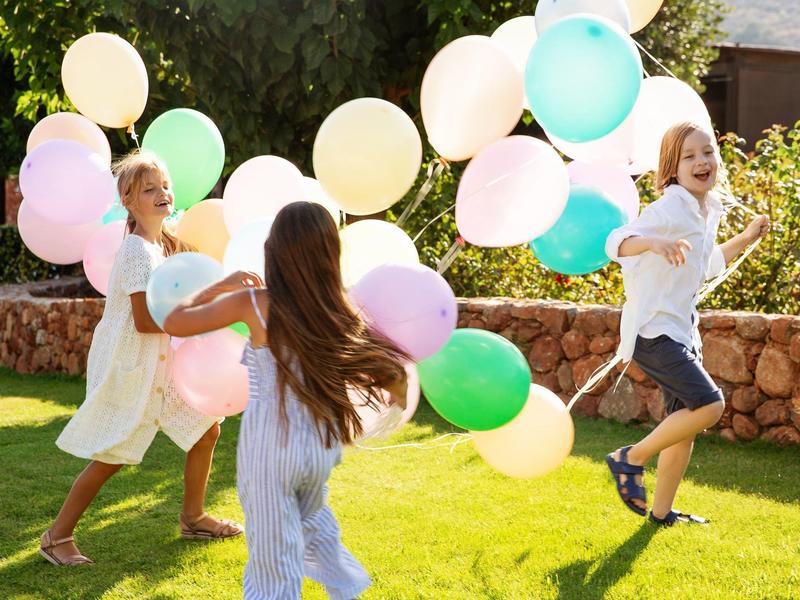 Three children running on grass holding colorful balloons in a sunny outdoor setting.