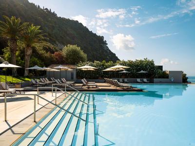 Infinity-Pool mit Liegestühlen und Sonnenschirmen neben einem bewaldeten Hang unter blauem Himmel.