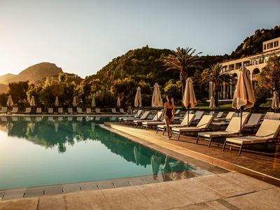 Ein ruhiger Hotelpool mit Sonnenliegen und Schirmen bei Sonnenuntergang vor bewaldeten Hügeln.