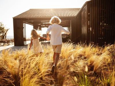 Zwei Kinder laufen durch hohes Gras zu einem modernen Holzhaus bei Sonnenuntergang.