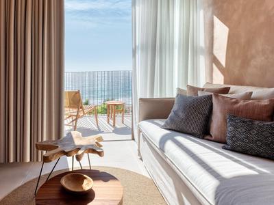 Sala moderna con sofá, cojines, silla de madera y vista al mar a través de grandes ventanas de vidrio.