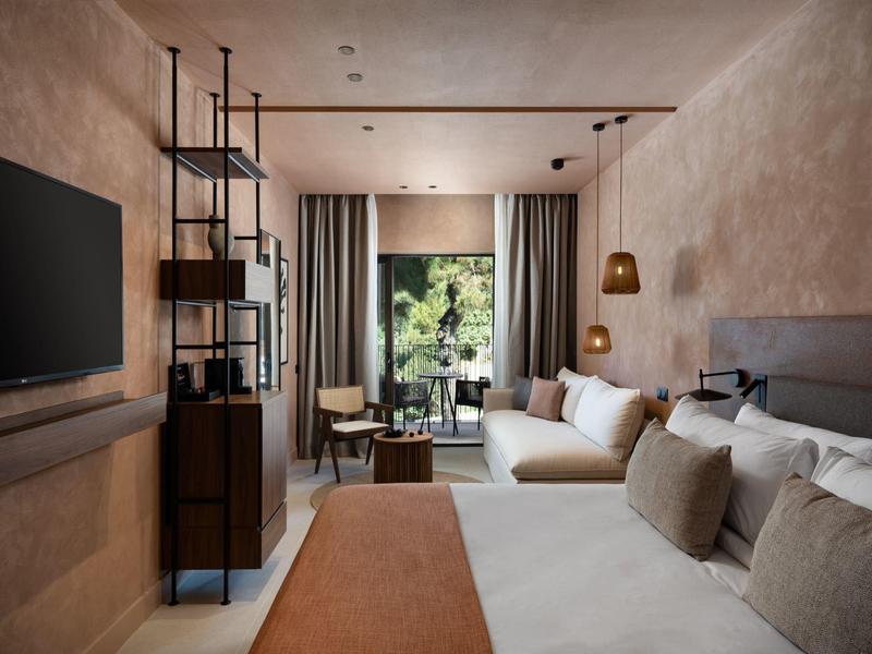 Acogedora habitación de hotel con cama grande, sofá, TV y vista a una terraza verde.