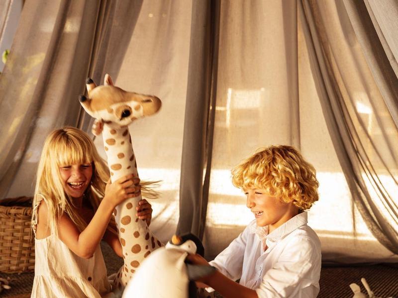 Niños juegan con animales de peluche frente a una cortina clara en una habitación acogedora.