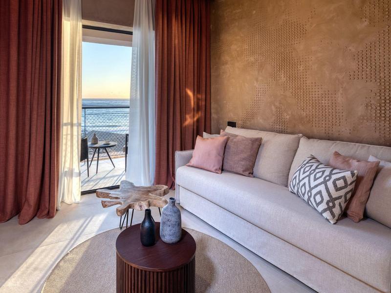 Sala acogedora con sofá, cojines y vista al mar a través de puertas abiertas.