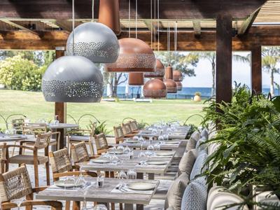 Área de restaurante abierta con mesas arregladas, lámparas modernas y vista al jardín y al mar.