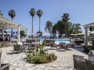 Hotelterrasse mit Sitzbereichen und Blick auf Pool und Palmen unter blauem Himmel.