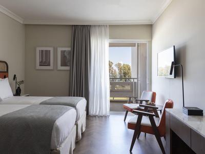 Habitación de hotel con dos camas individuales, sillas, TV y balcón con vista al exterior.