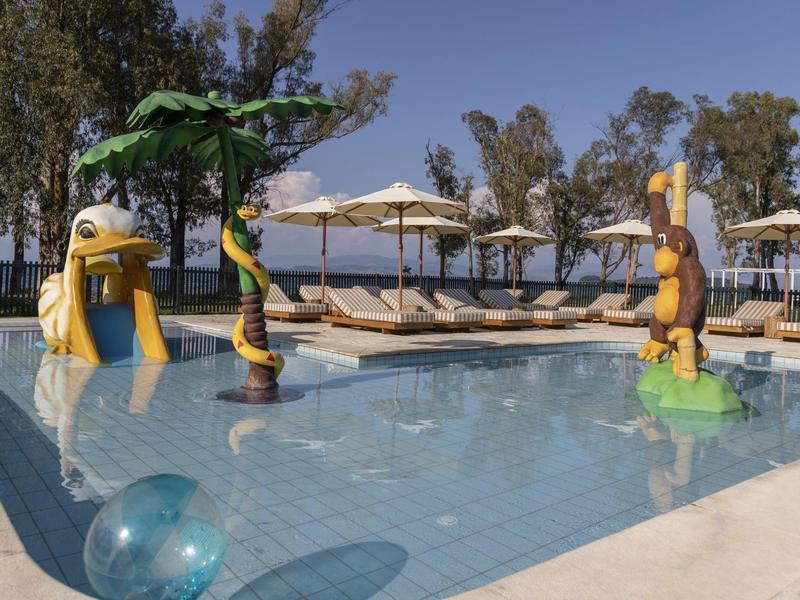 Piscina infantil con toboganes de agua, sombrillas y tumbonas en un área exterior con árboles.