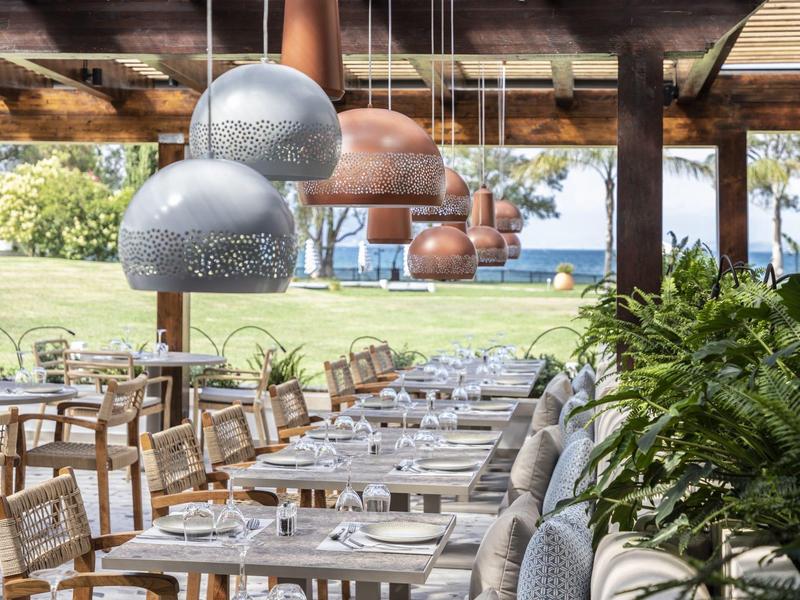 Area esterna moderna di un ristorante con tavoli, sedie e lampade sospese che si affacciano su un prato verde.